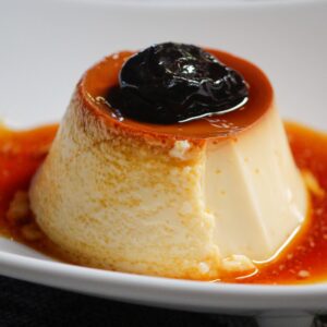 Creamy flan dessert topped with caramel sauce and a prune on a white plate.